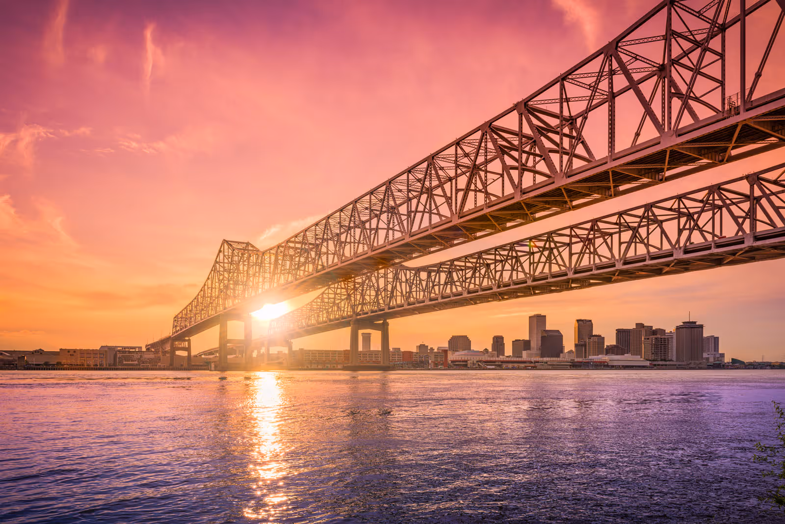 Steel cantilever bridge over river at sunset with city skyline in the background.