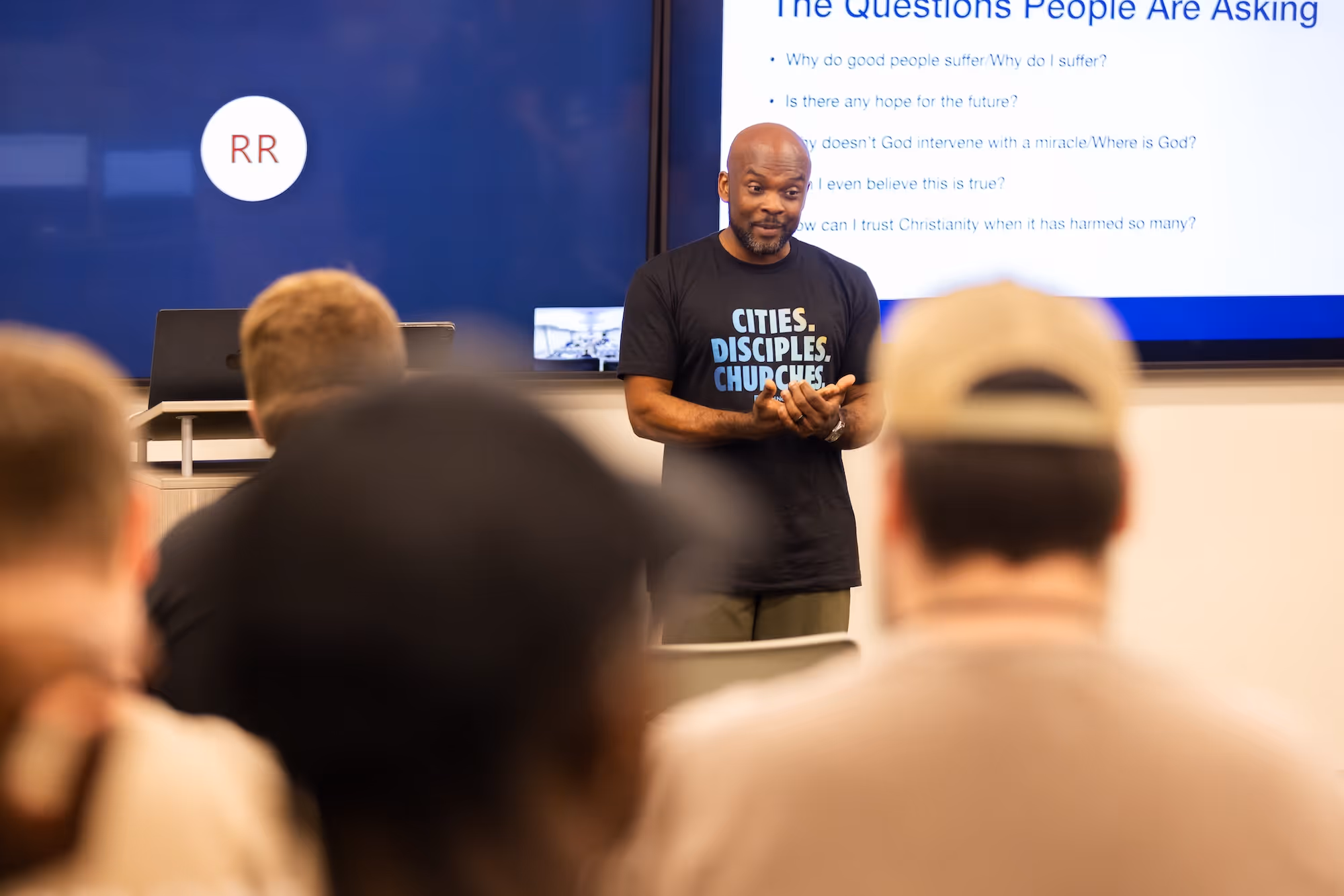 Man in black T-shirt giving a presentation to an audience with text displayed on a screen behind him.