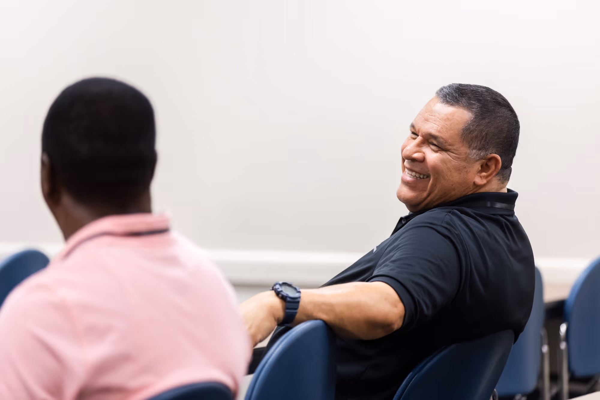 Man in black shirt smiling and leaning back in a chair during a conversation with another man in a pink shirt.
