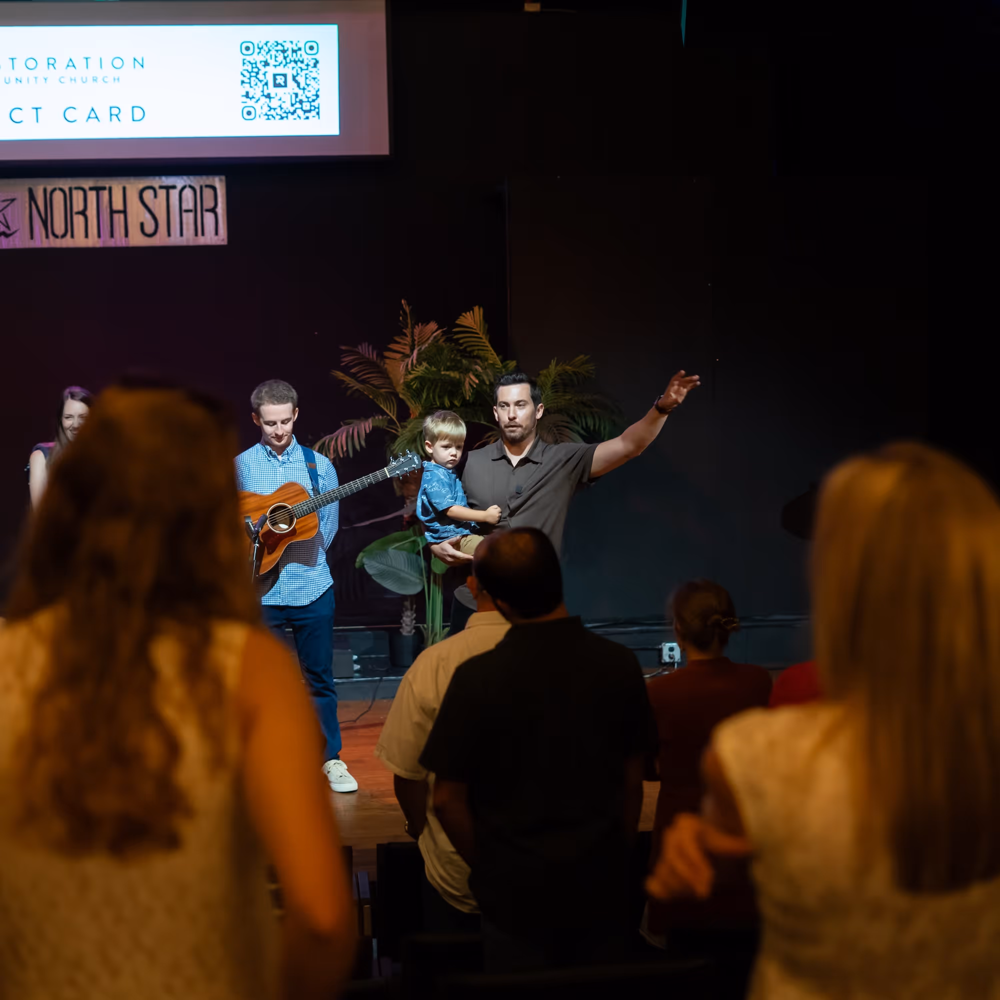 Man holding a child with raised arm speaking to a seated audience while another man stands with a guitar.
