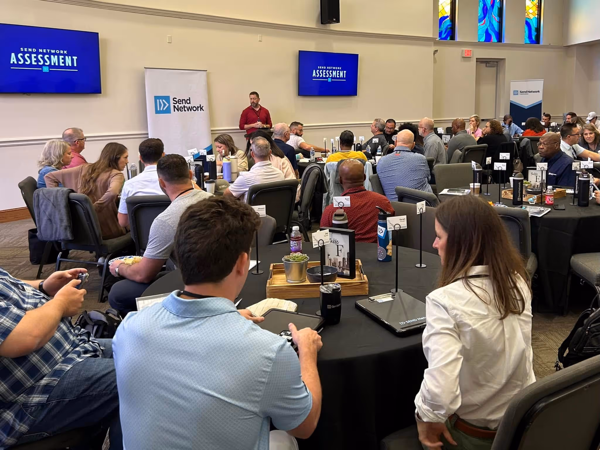 People seated around tables in a conference room attending a Send Network Assessment presentation.