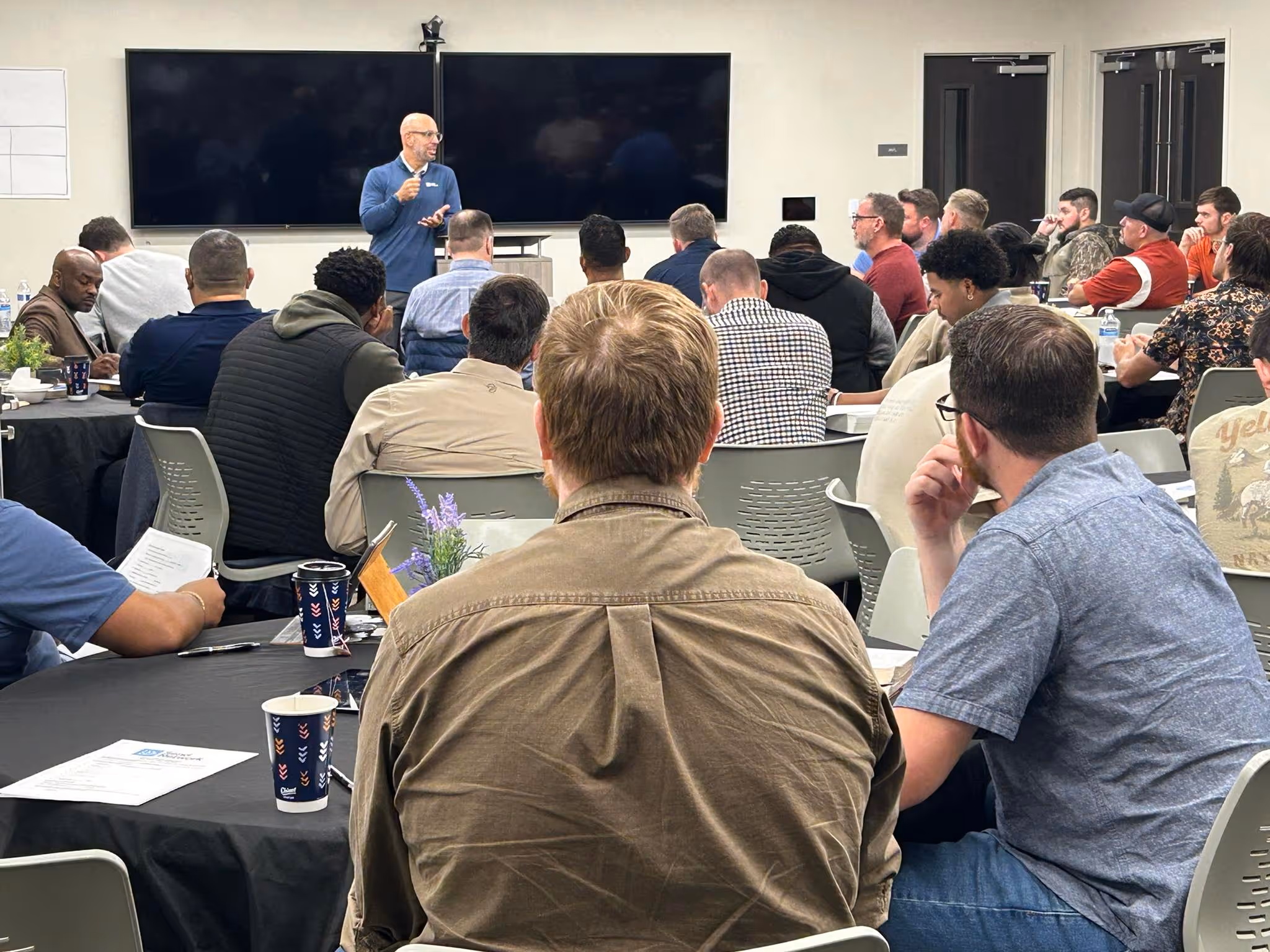 A man in a blue shirt speaks in front of an audience seated at round tables in a conference room.
