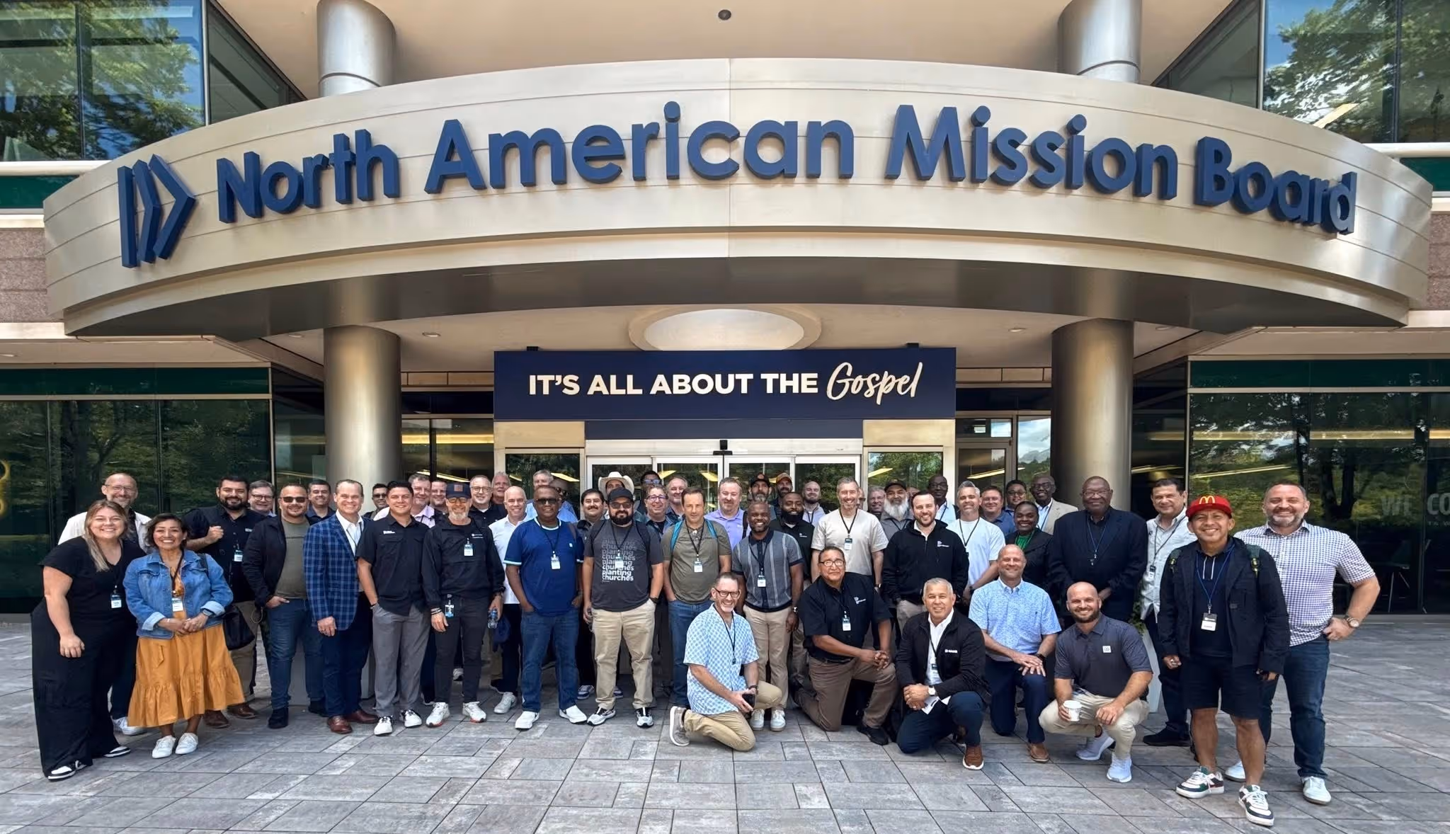 Group of men and women posing in front of North American Mission Board building with gospel sign above entrance.