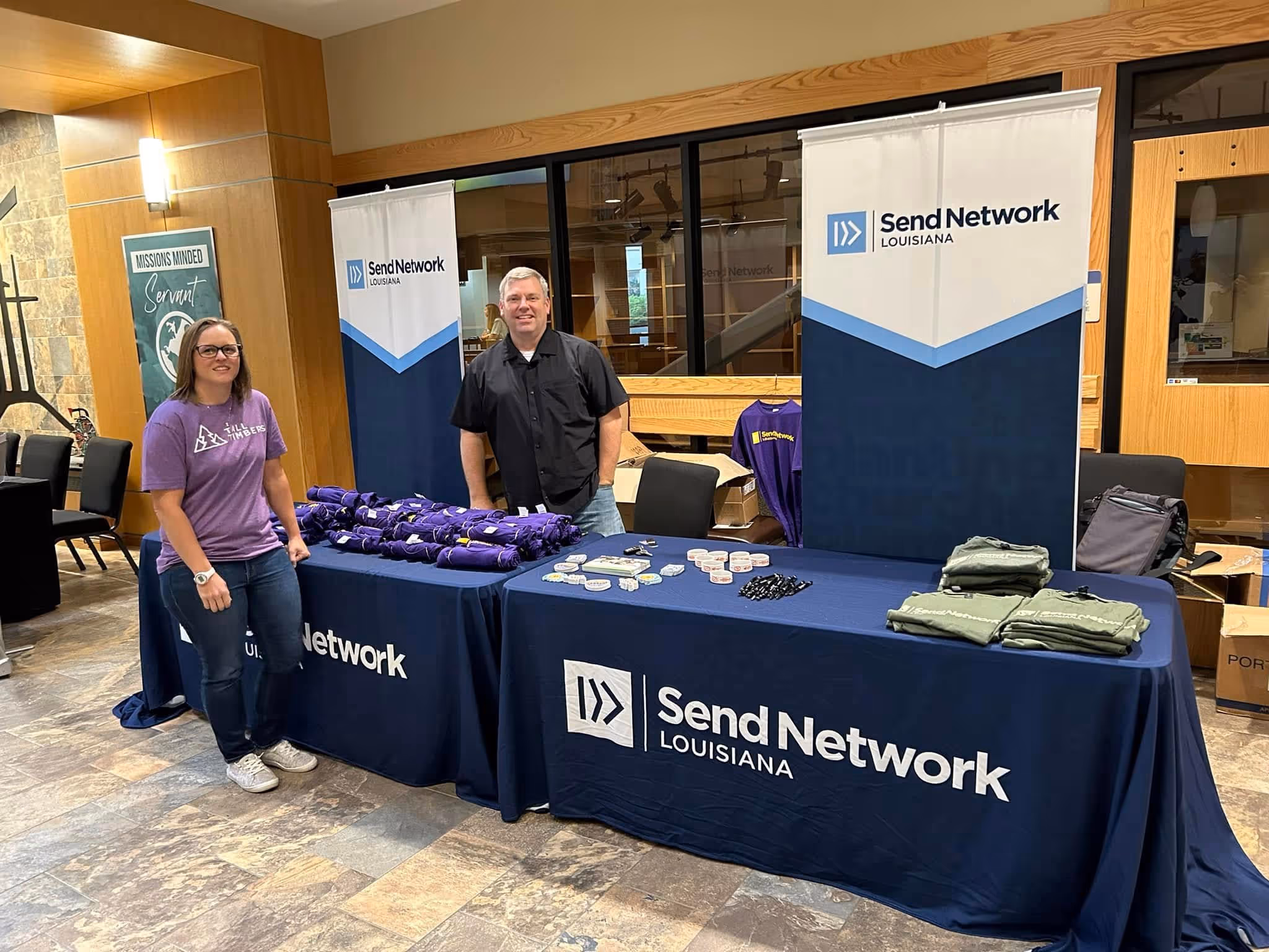 Two people standing behind Send Network Louisiana tables with branded shirts and promotional items in a lobby area.