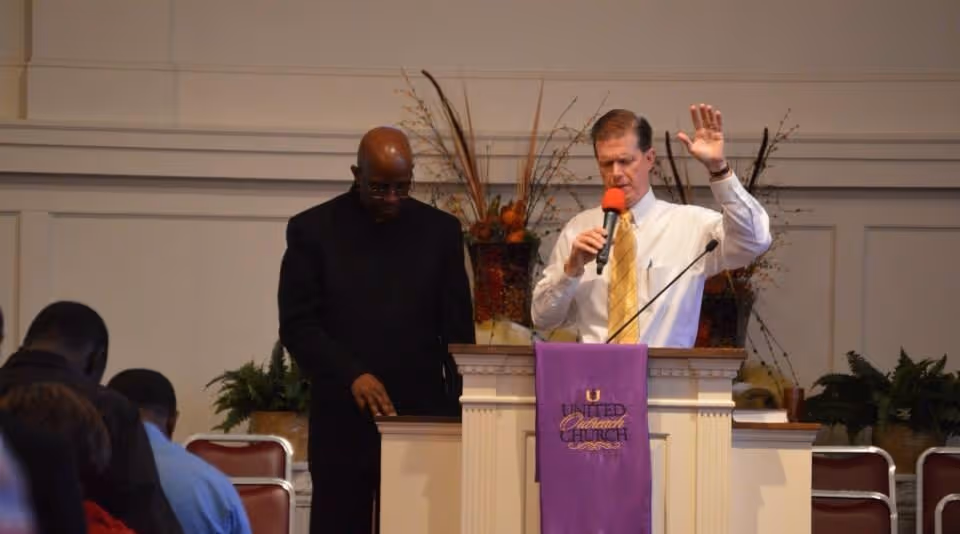 Two men standing at a church pulpit during a service, one praying and one speaking into a microphone with hand raised.
