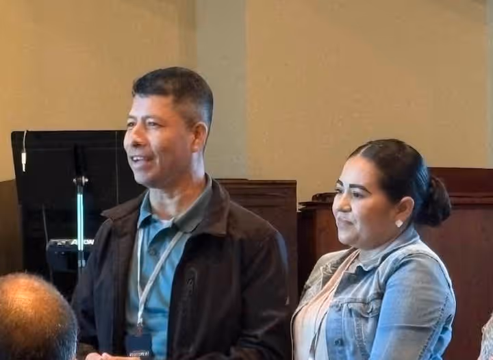 Man and woman standing indoors, speaking or presenting to a seated audience.