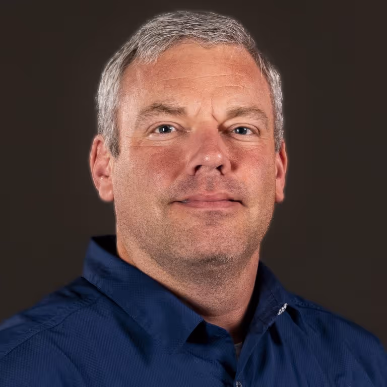 Man with short gray hair wearing a blue collared shirt against a dark background.