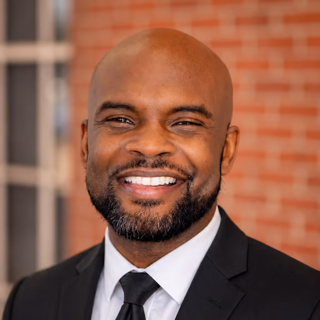 Smiling bald man with a beard wearing a black suit, white shirt, and black tie in front of a brick wall.
