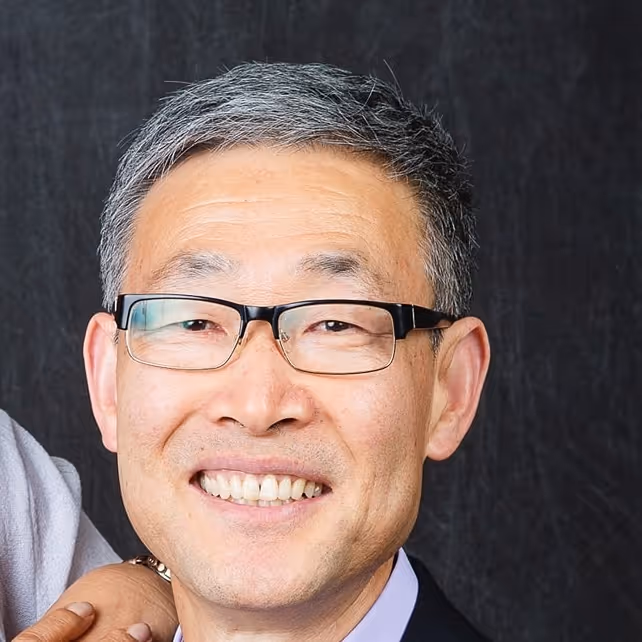 Smiling Asian man with gray hair and glasses against a dark background.