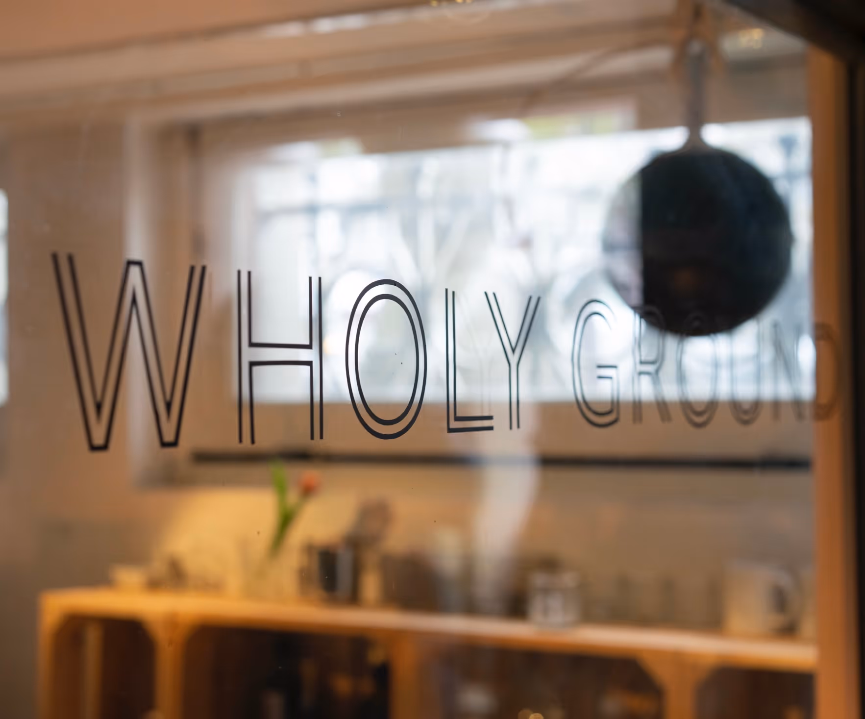 Glasdeur met de tekst 'WHOLY GROUND' en een wazig interieur met meubels en een plant erachter.