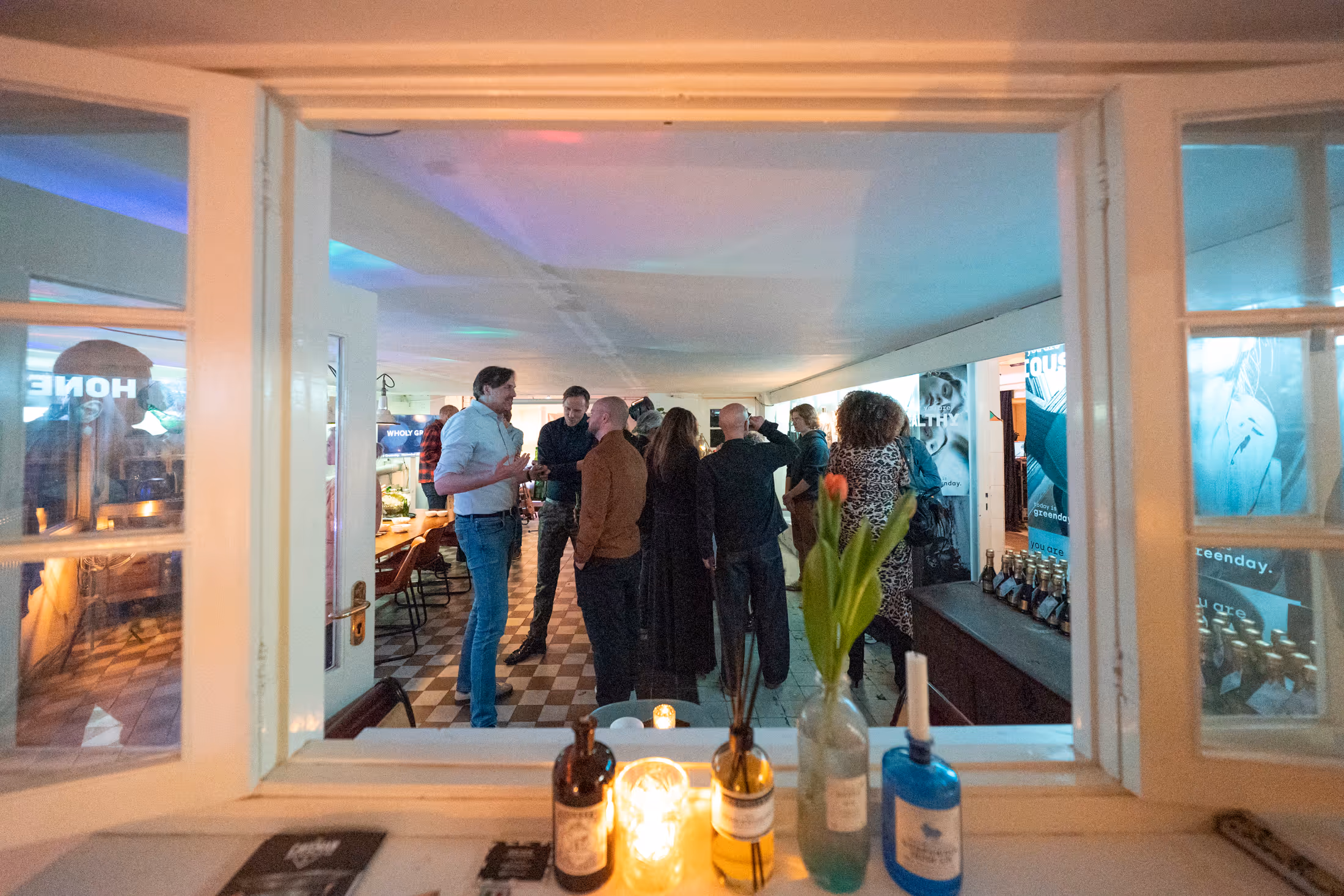 Groep mensen die in een helder verlichte kamer staan en met elkaar praten, gezien door een open raam met flessen en een bloem op de vensterbank.