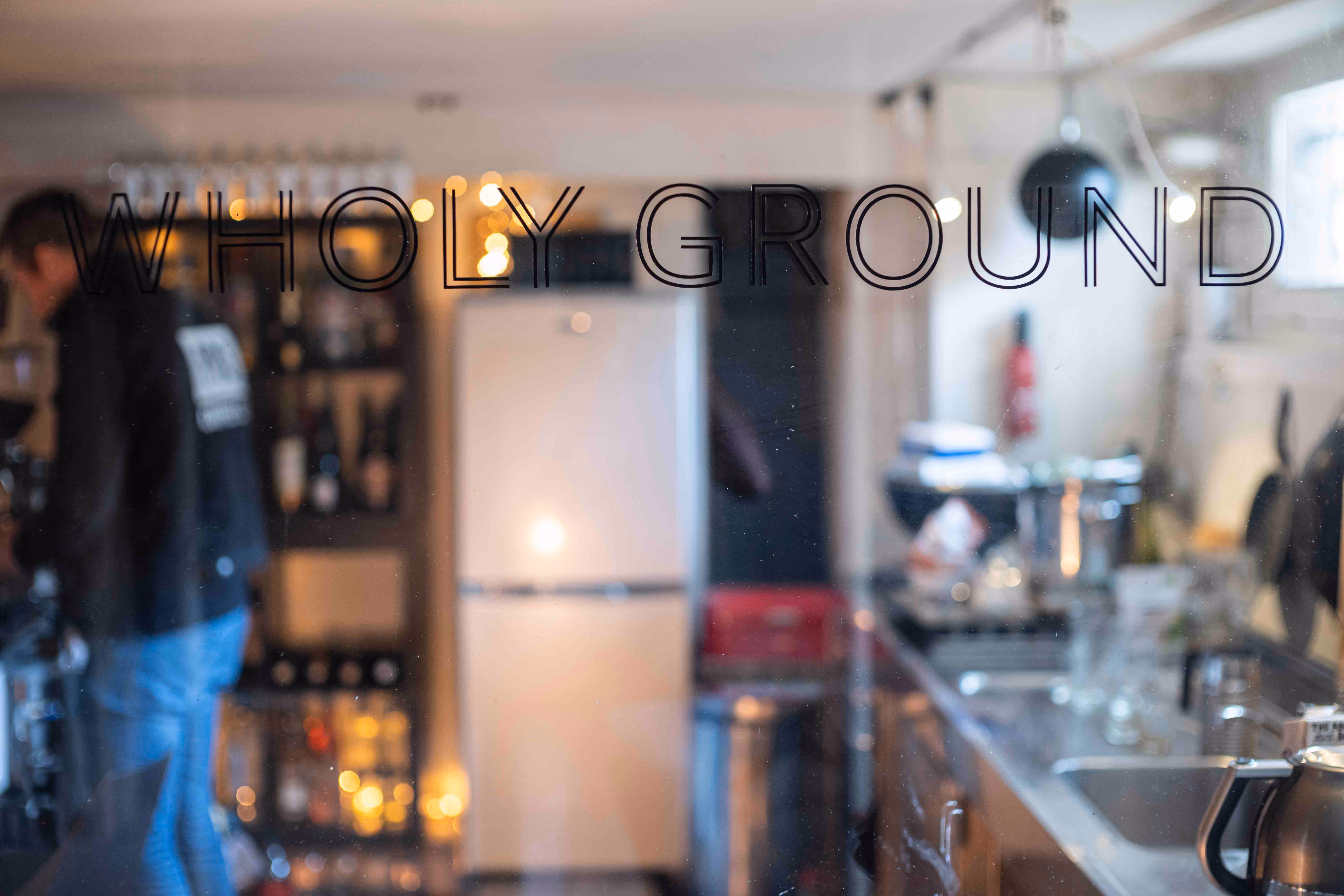 Door met de tekst 'WHOLY GROUND' met daarin vaag een keuken en een persoon in een zwarte jas zichtbaar.