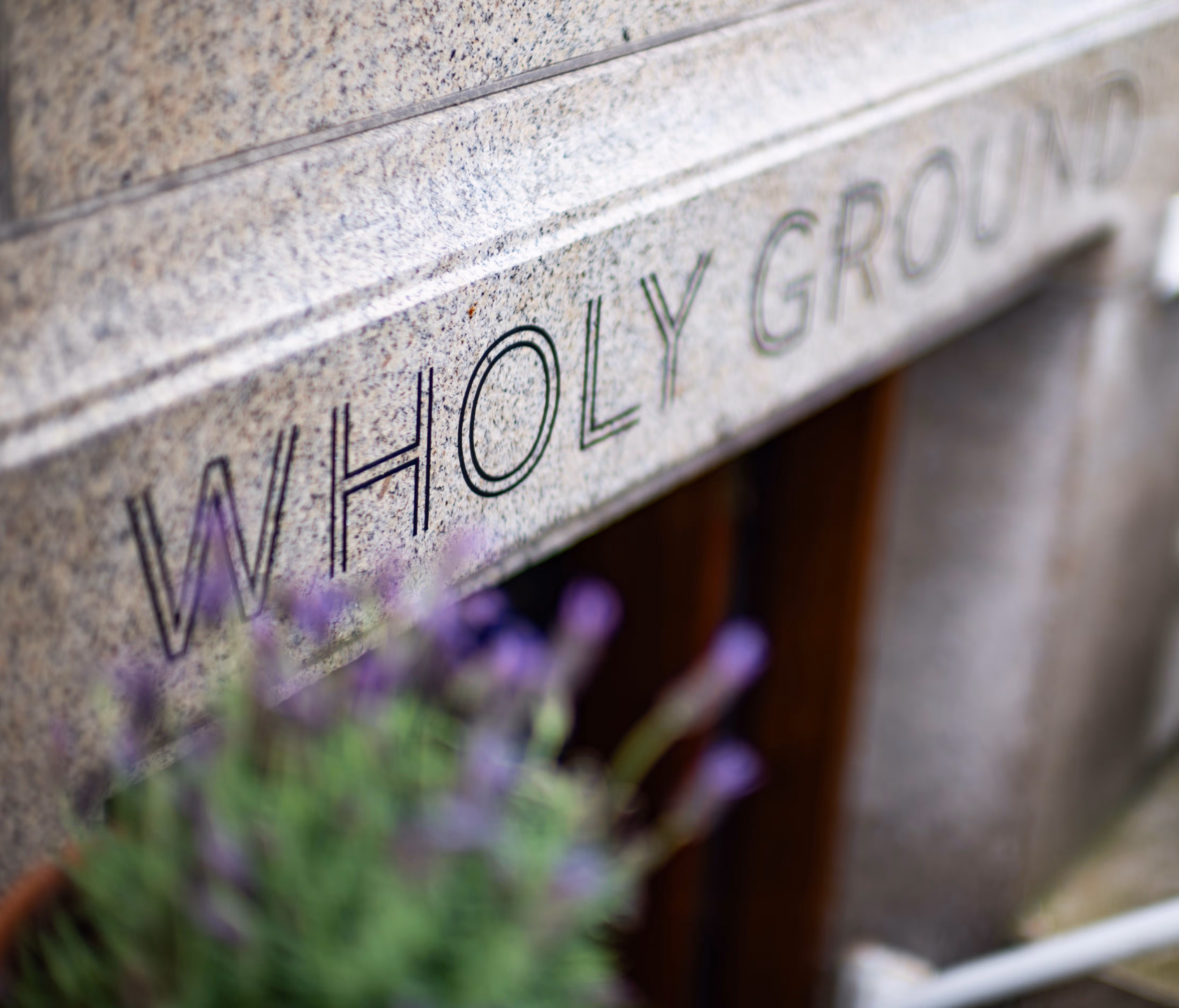 Grijze stenen gevel met het woord 'WHOLELY GROUND' gegraveerd, wazige paarse bloemen op de voorgrond.