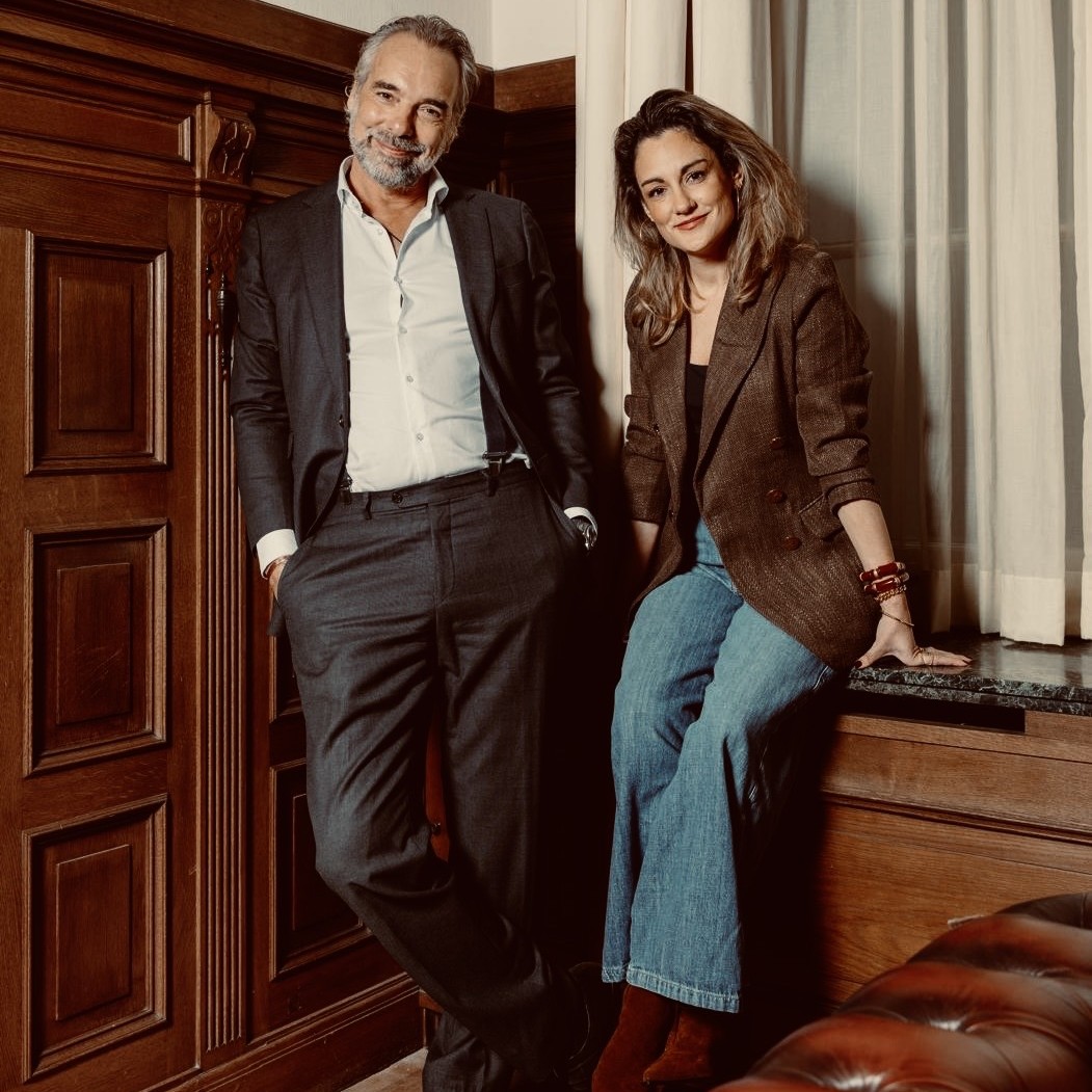 Man in pak en vrouw in blazer en wijde jeans zitten ontspannen in een kamer met houten lambrisering en gordijnen.