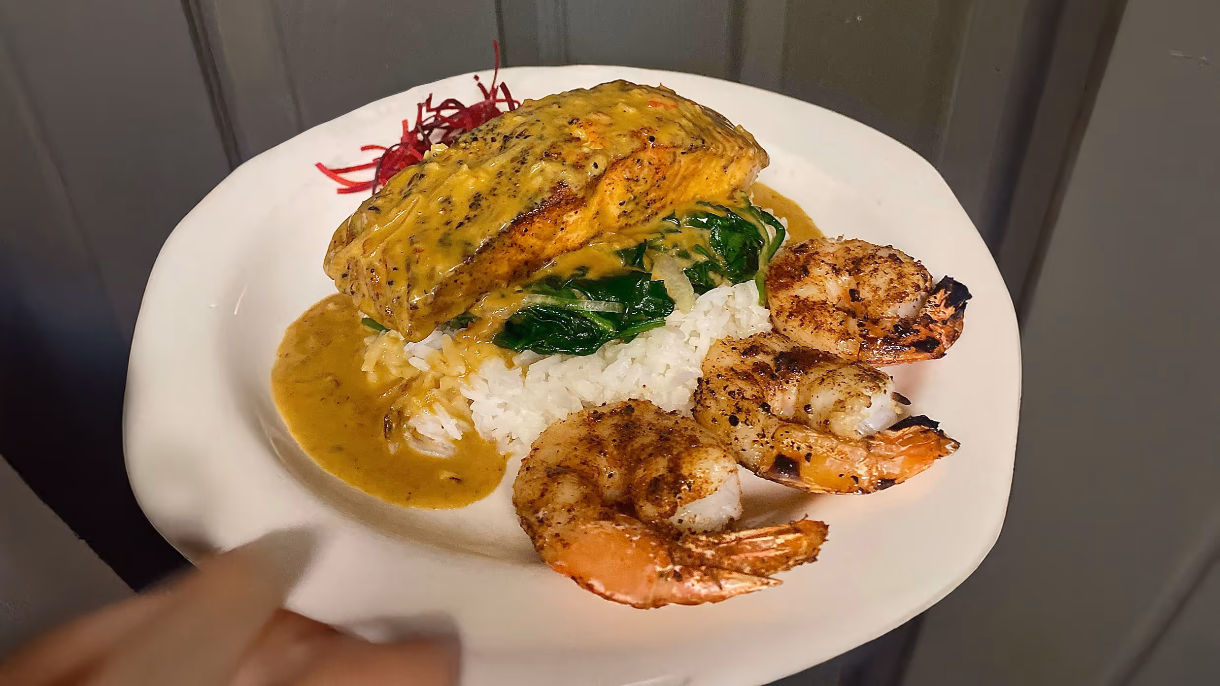 A plate of grilled salmon on rice with creamy sauce, sautéed spinach, and three grilled shrimp, garnished with shredded red beets.