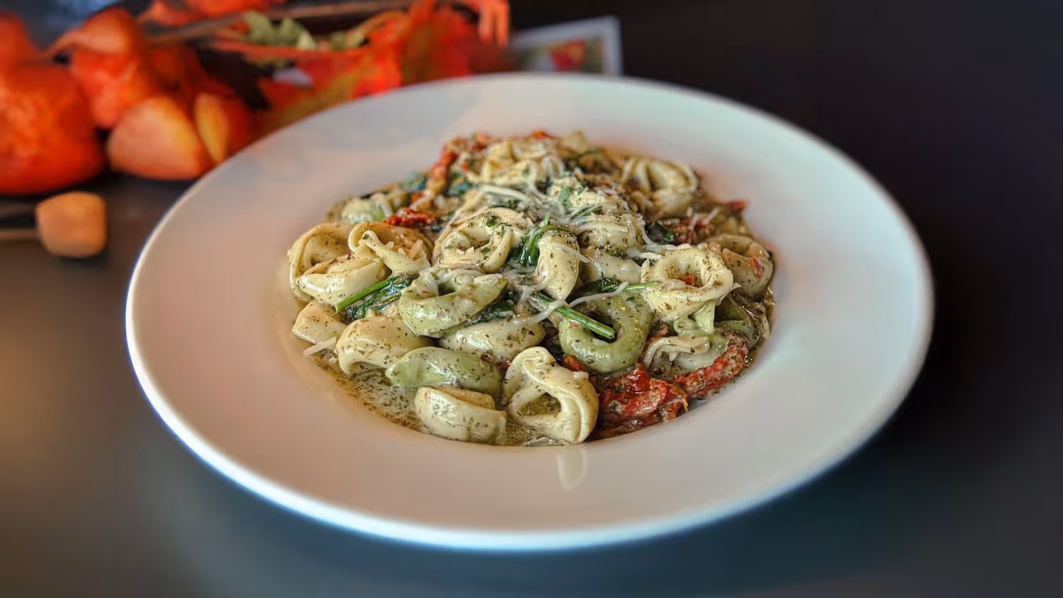 A plate of tortellini pasta in a creamy pesto sauce, garnished with sun-dried tomatoes and herbs, sits on a dark table.