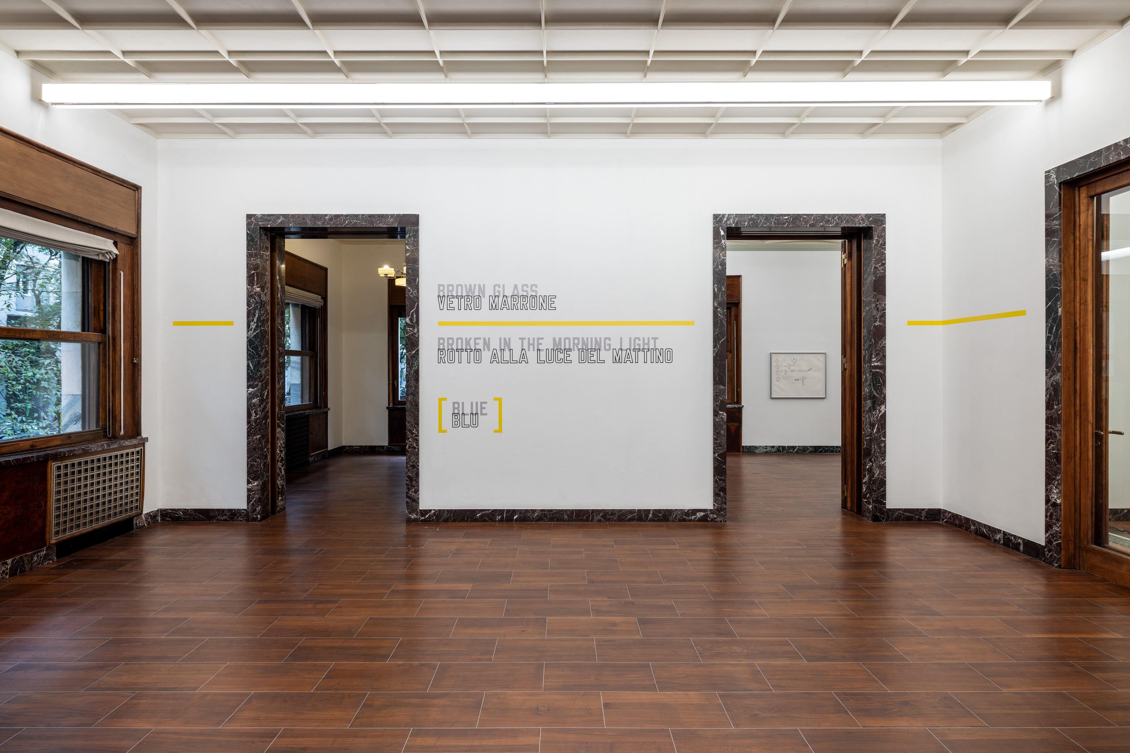 BROWN GLASS sits between two doorways, with lengths of yellow continuing beyond the verbal group on either side. Image Credit: Installation view. Courtesy of the Artist and MASSIMODECARLO. Photo: Roberto Marossi.