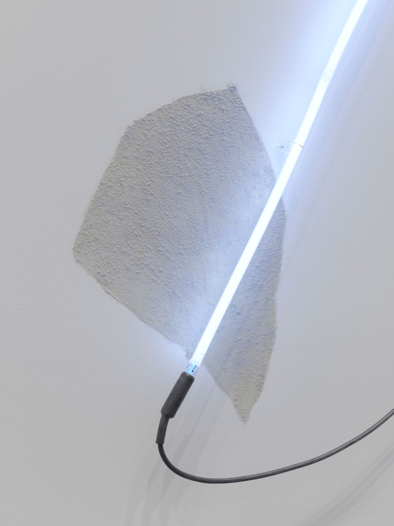 The lower cap rests over the sheet's torn edge. The surface is pebbled; a clear standoff holds the tube off the wall. Image Credit: Arced Neon (Cool White), 1968 (detail). Courtesy Estate of Keith Sonnier and David Kordansky Gallery. Photo: Jeff McLane.