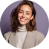 Smiling woman with light brown hair wearing a beige turtleneck and blazer against a purple background.