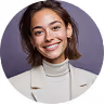 Smiling woman with shoulder-length brown hair wearing a light turtleneck and beige blazer against a gray background.