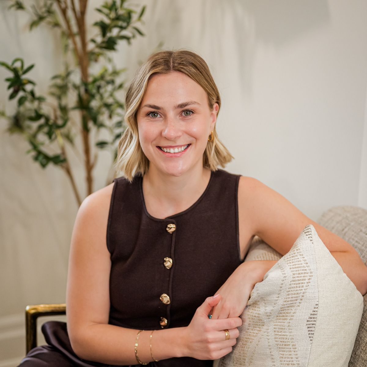 Smiling woman with short blonde hair wearing a sleeveless black top sitting on a couch with a cushion.