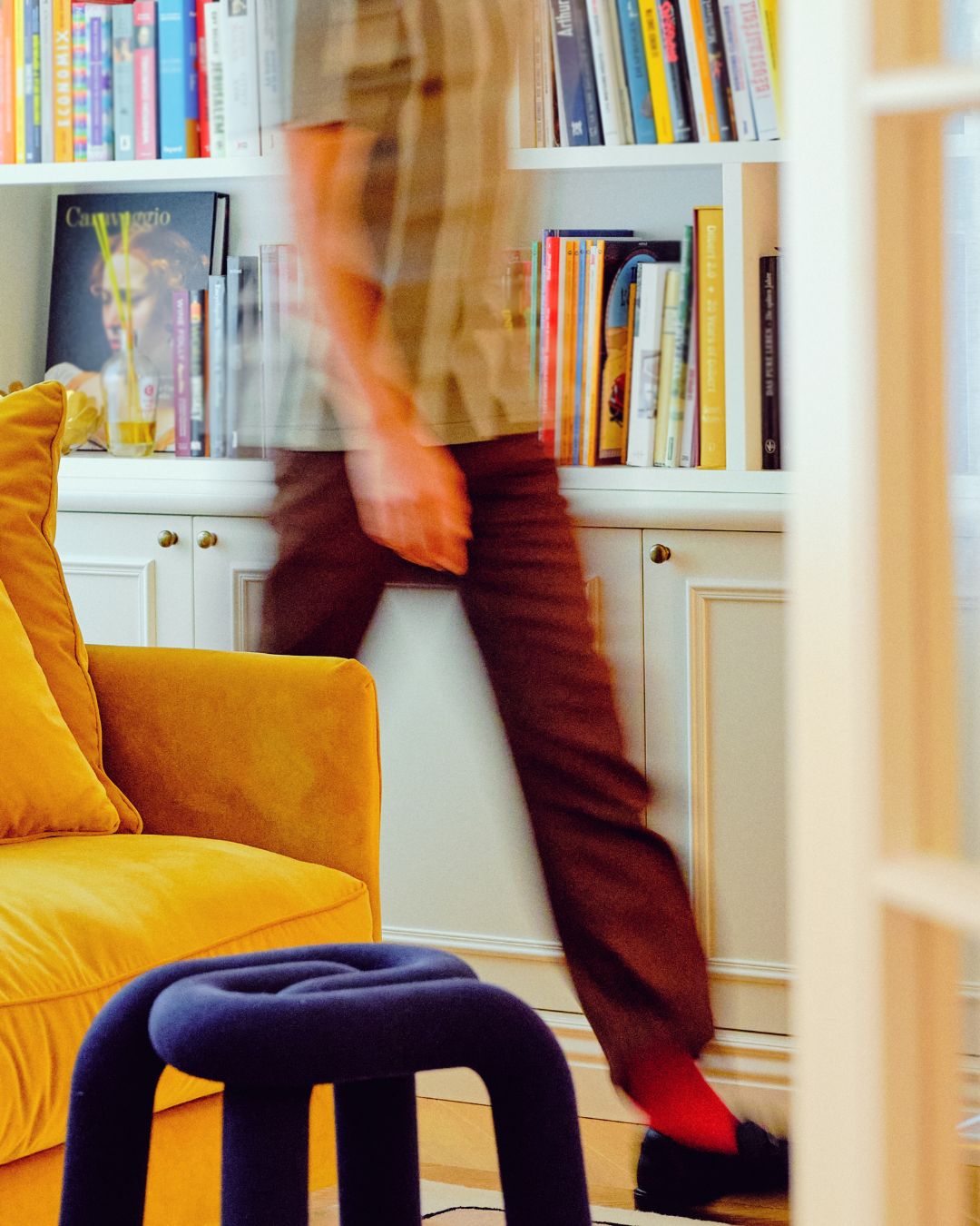 Blurred person walking past a yellow sofa and bookshelf filled with books in a cozy living room.