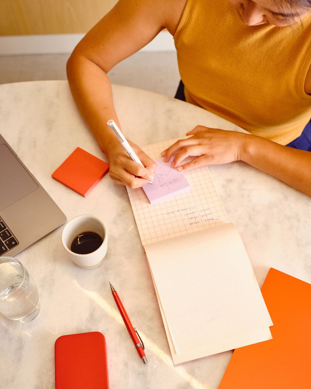 Person in yellow top writing on purple sticky notes over a notebook on a marble table with coffee and laptop.