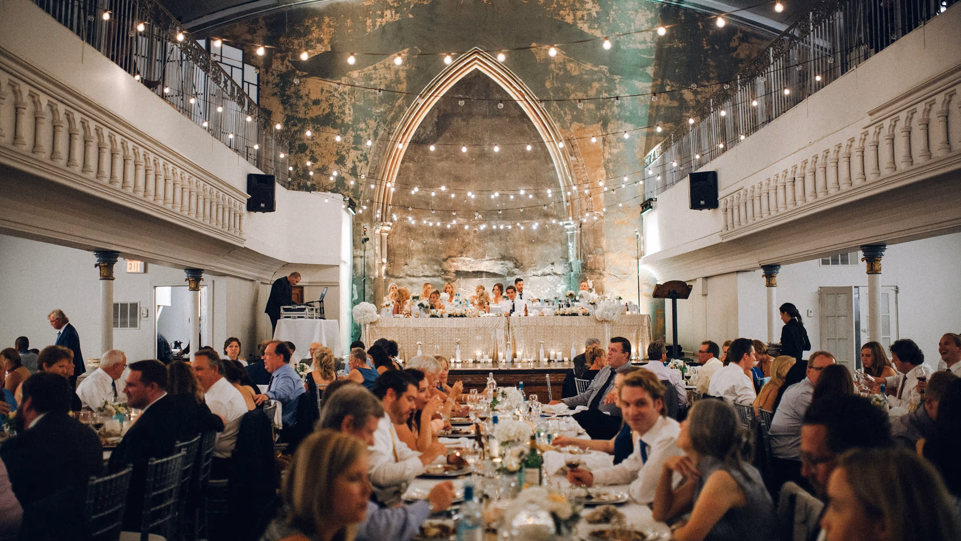 The Alcmy - Wedding Serve - Wedding reception in a large hall with guests seated at long tables, string lights overhead, and the bridal party at a decorated head table on a stage.
