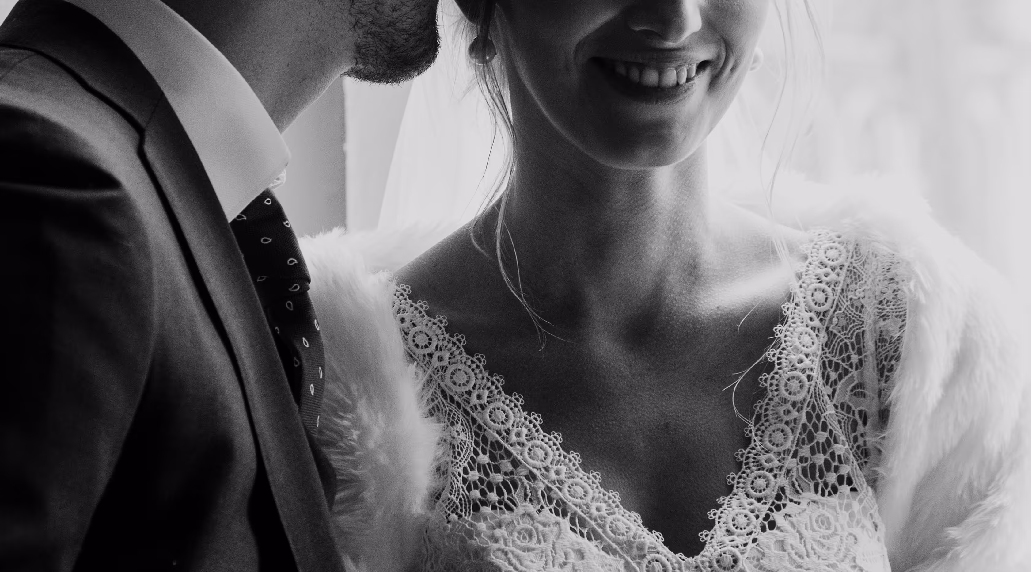 The Alcmy - Weddings - Black and white close-up of a bride smiling in a lace wedding dress and a groom in a suit and patterned tie.