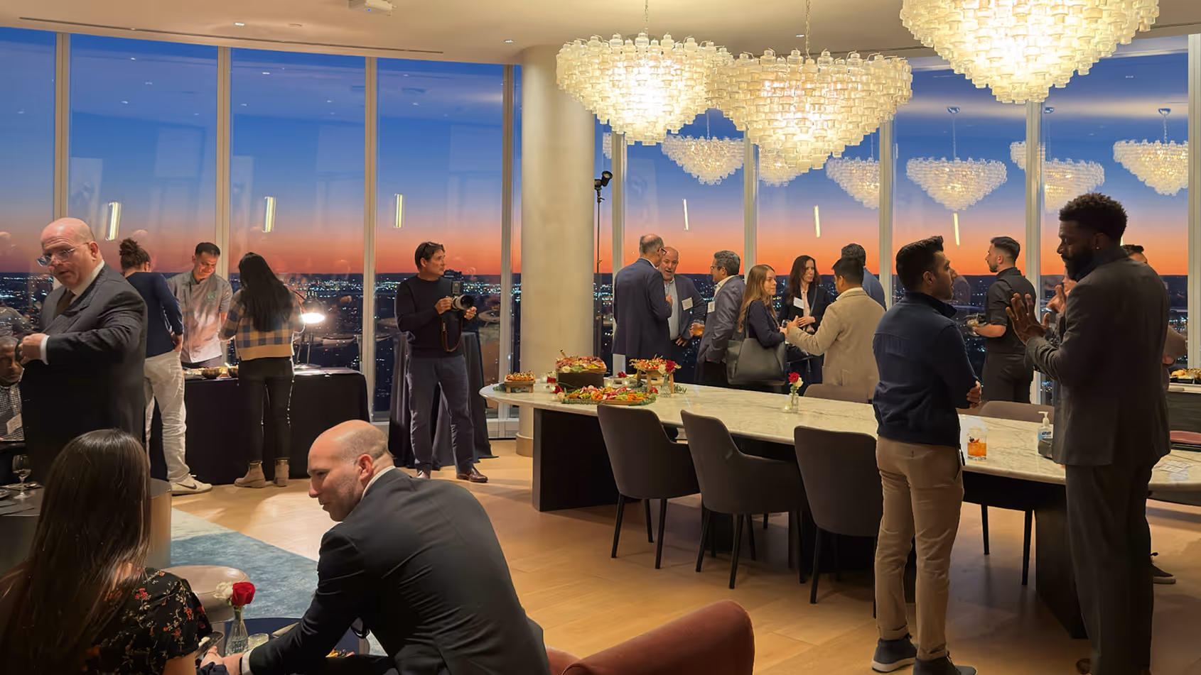 People socializing in a modern lounge with large floor-to-ceiling windows showing a colorful sunset cityscape outside.