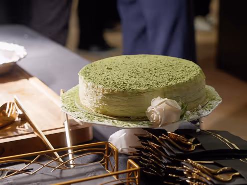 Round green tea cake with powdered topping and a white rose decoration on a table with golden cutlery.