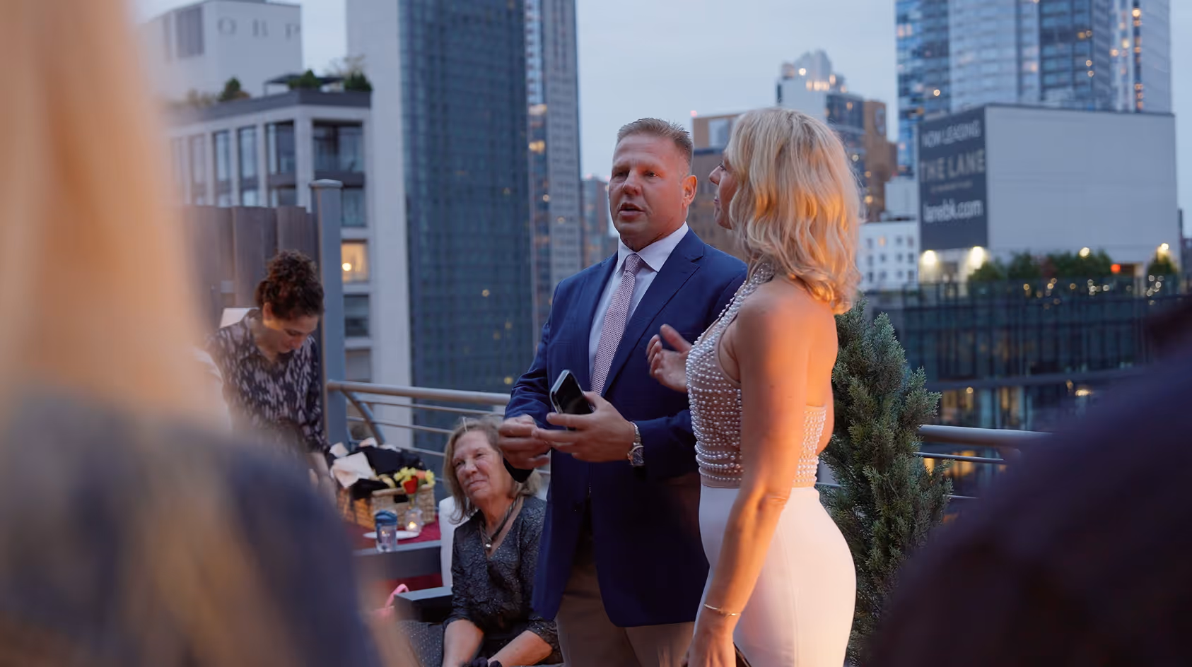 Man in suit holding a phone speaks next to a woman in a white dress on a rooftop with city buildings in the background during dusk.
