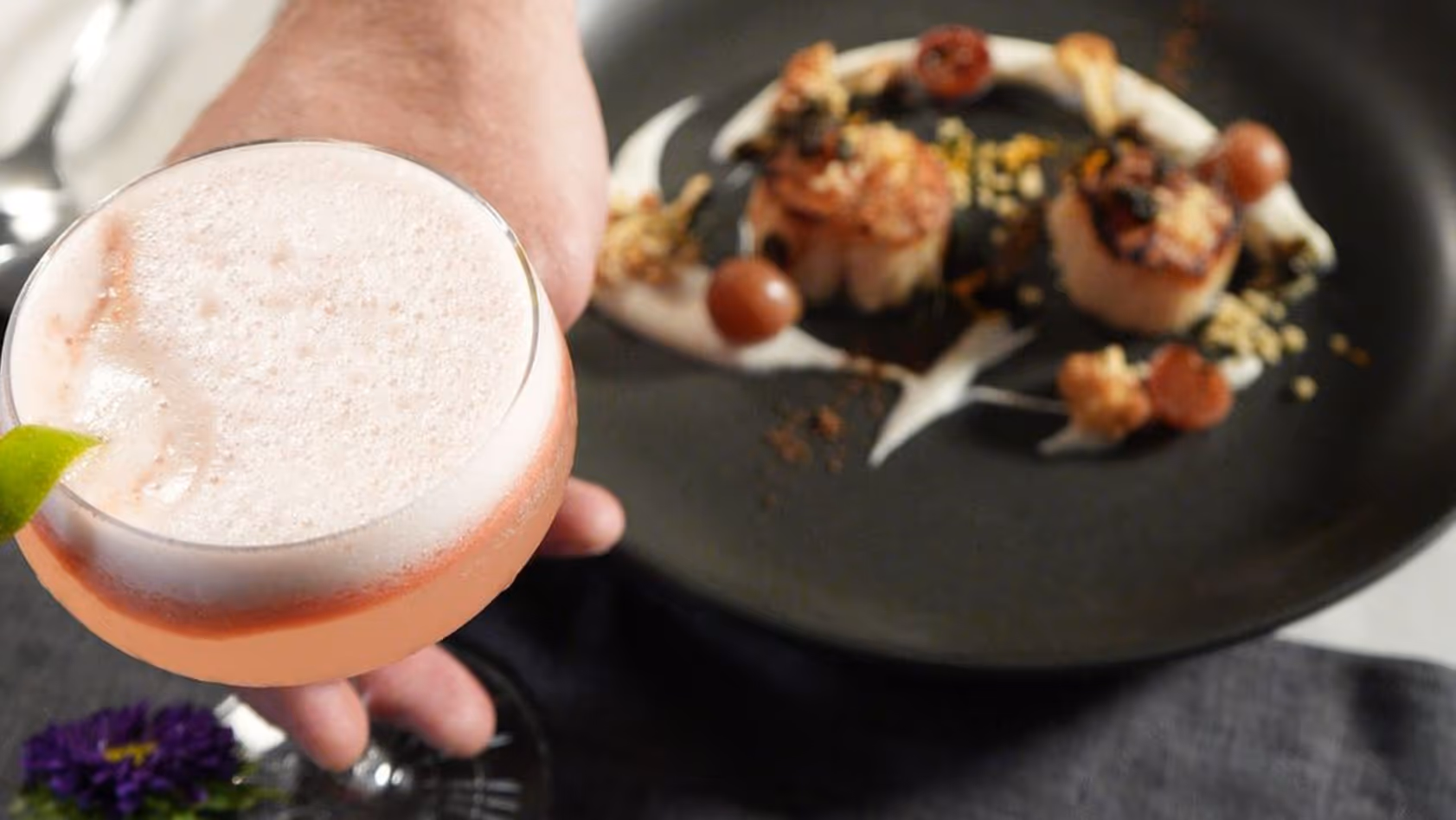 Hand holding a pink frothy cocktail garnished with a lime wedge, with a black plate of seared scallops and garnish in the background.