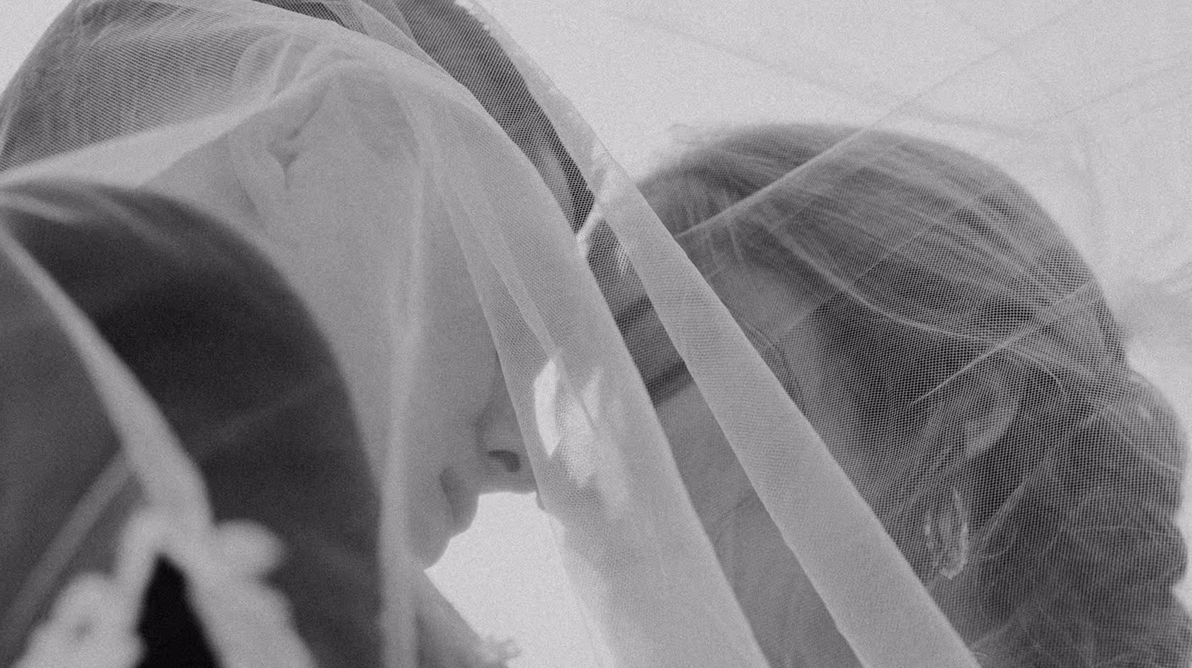 Black and white close-up of a bride and groom touching foreheads under a sheer wedding veil.