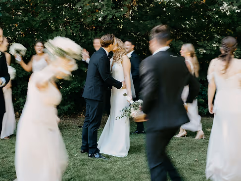 Bride and groom kissing outdoors surrounded by blurred bridesmaids and groomsmen in formal attire.