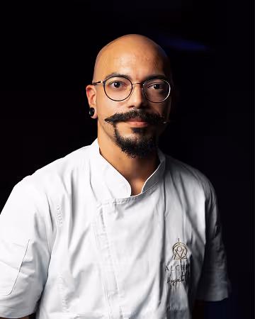 Bald man with glasses, a handlebar mustache, and earrings, wearing a white chef's coat against a black background.
