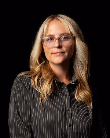 Woman with long blonde hair wearing clear-framed glasses and a black pinstripe shirt against a black background.