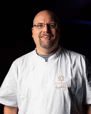 Smiling bald man with glasses wearing a white chef's coat with Alchemy Hospitality logo on black background.