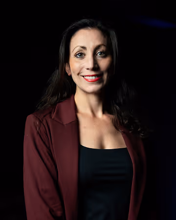 Smiling woman with long dark hair wearing a black top and maroon blazer against a black background.