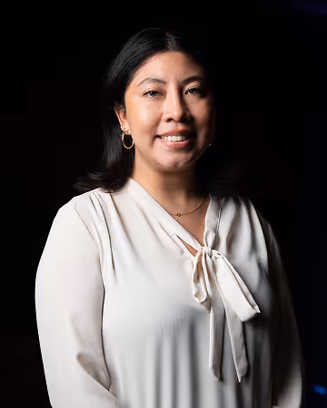 Smiling woman with dark hair wearing a white blouse with a bow tie detail and gold hoop earrings against a black background.