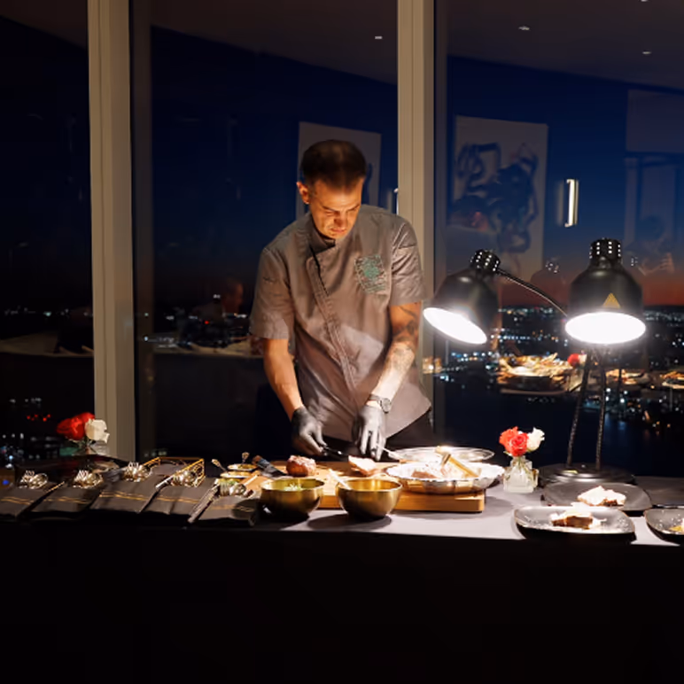 Chef wearing gloves preparing food on a table illuminated by two lamps with a nighttime cityscape in the background.
