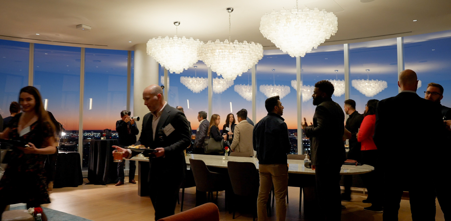 People socializing indoors during sunset, with large glass windows and heart-shaped chandeliers overhead.