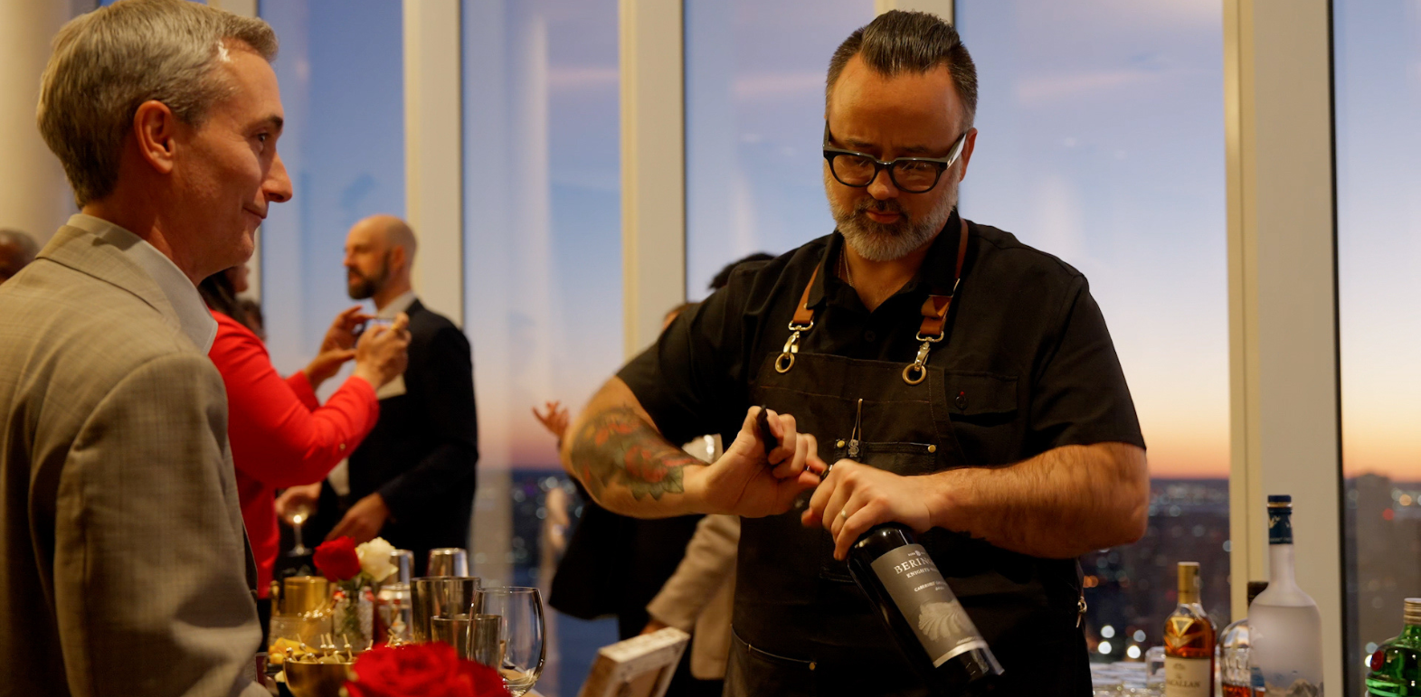 Bartender with tattoos and glasses opening a bottle of wine at a crowded evening event with large windows showing a cityscape at sunset.