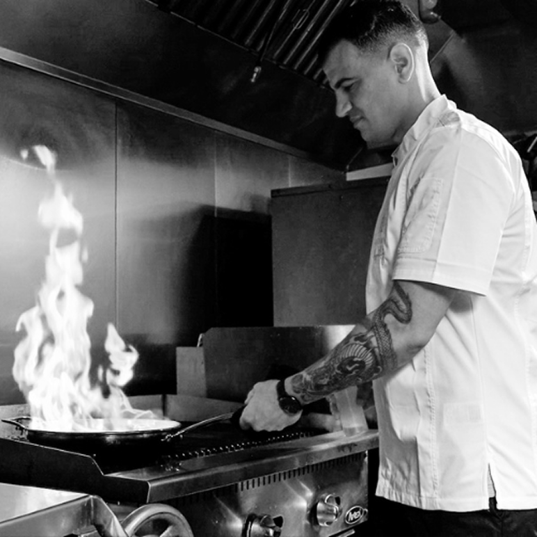 Chef with a tattoo on his arm cooking on a stove with flames rising from a pan in a professional kitchen.