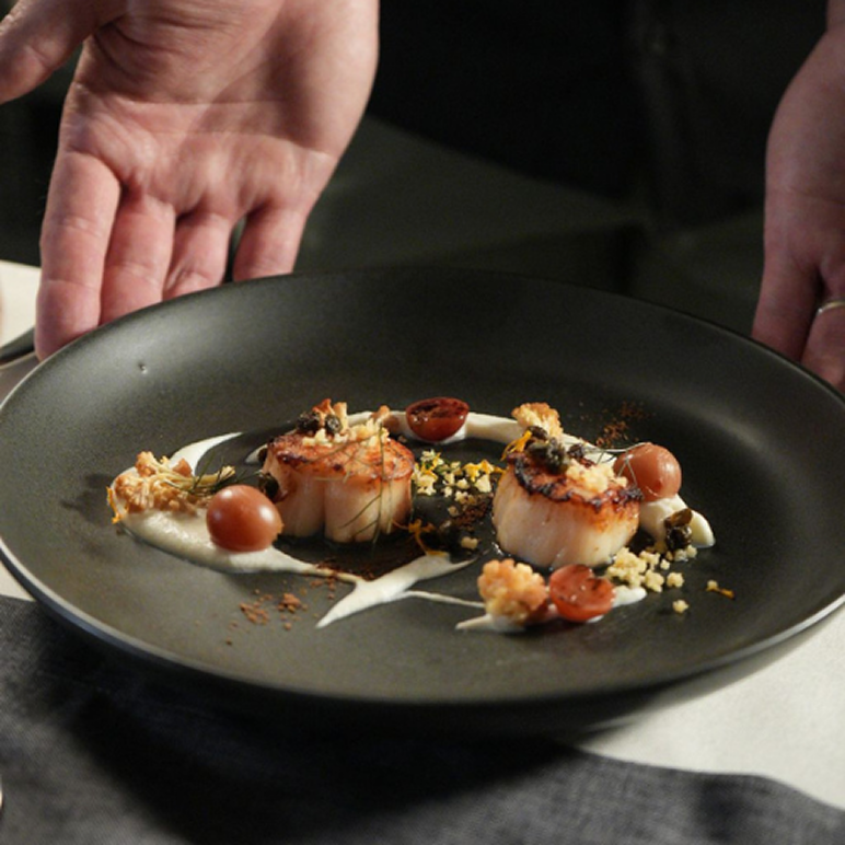 Two seared scallops garnished with small tomatoes, herbs, and cream sauce on a black plate held by two hands.