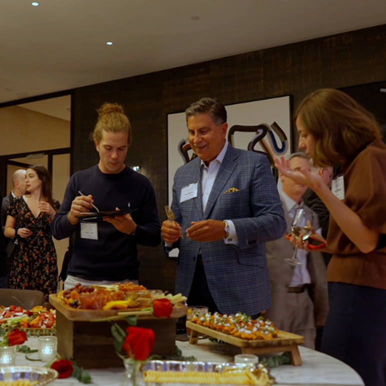 People enjoying appetizers and drinks at a social gathering with a table full of food and red roses.