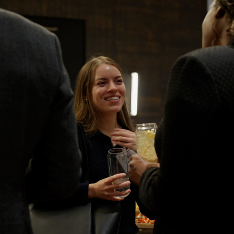 Smiling woman holding a glass and talking with two people in a dimly lit gathering.