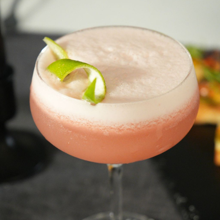 Pink cocktail with frothy top garnished with a twisted lime peel in a coupe glass.