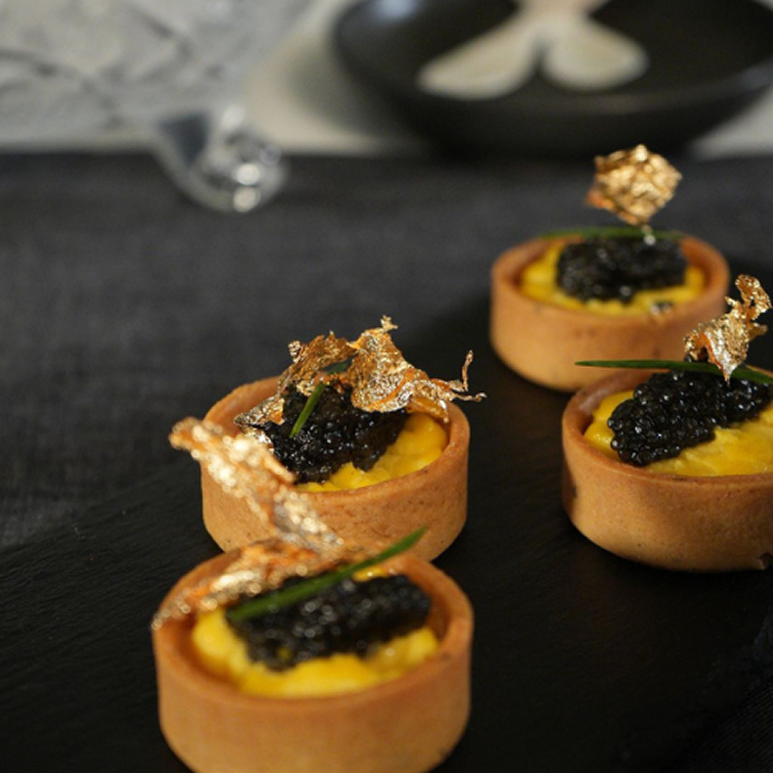 Four elegant tartlets with yellow filling topped with black caviar and decorative gold leaf on a black serving board.