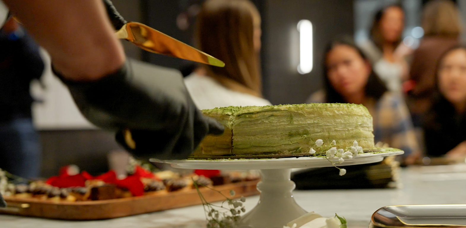 Person wearing black gloves slicing a green layered cake with a knife at a gathering.